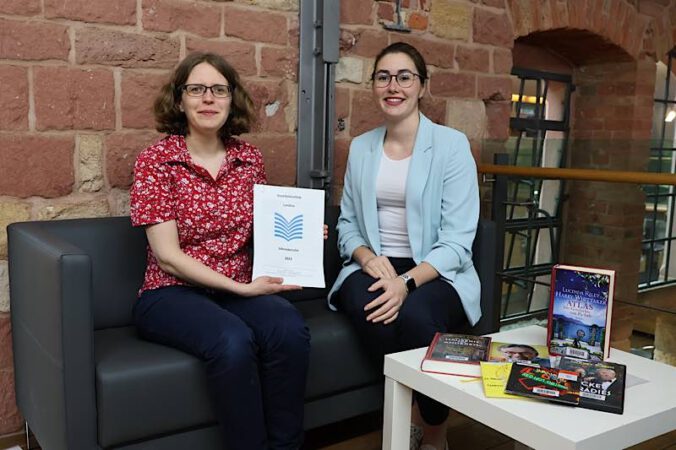 Bibliotheksleiterin Amelie Löhlein (l.) und Beigeordnete Lena Dürphold mit dem Jahresbericht 2023 und einer Auswahl der meistausgeliehenen Medien aus 2023 (Quelle: Stadt Landau)